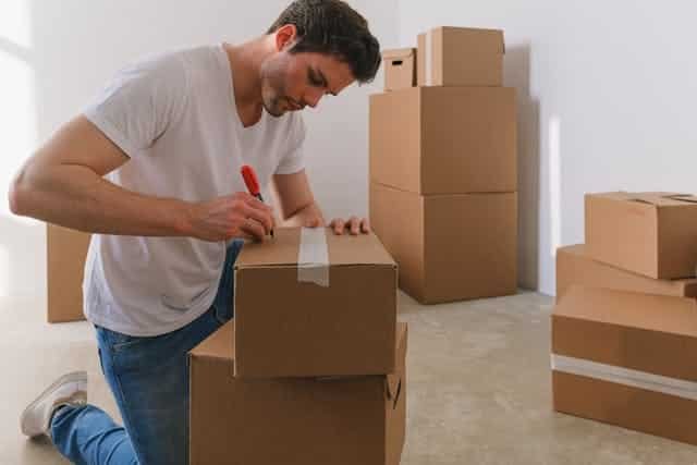 A man in a white shirt labeling cardboard boxes on the floor.